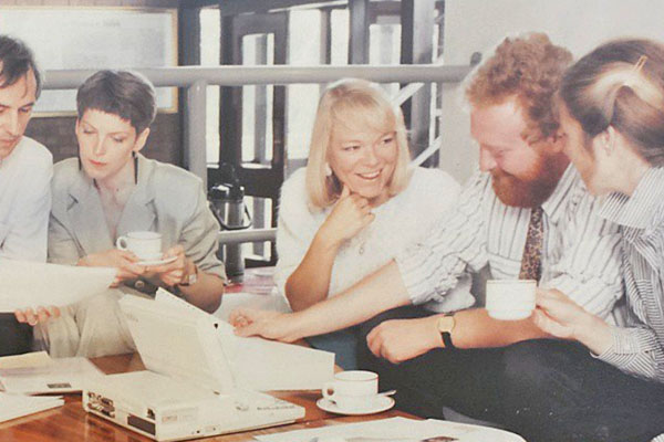 Five students sat around a table conversing with cups of tea whilst looking at documents.